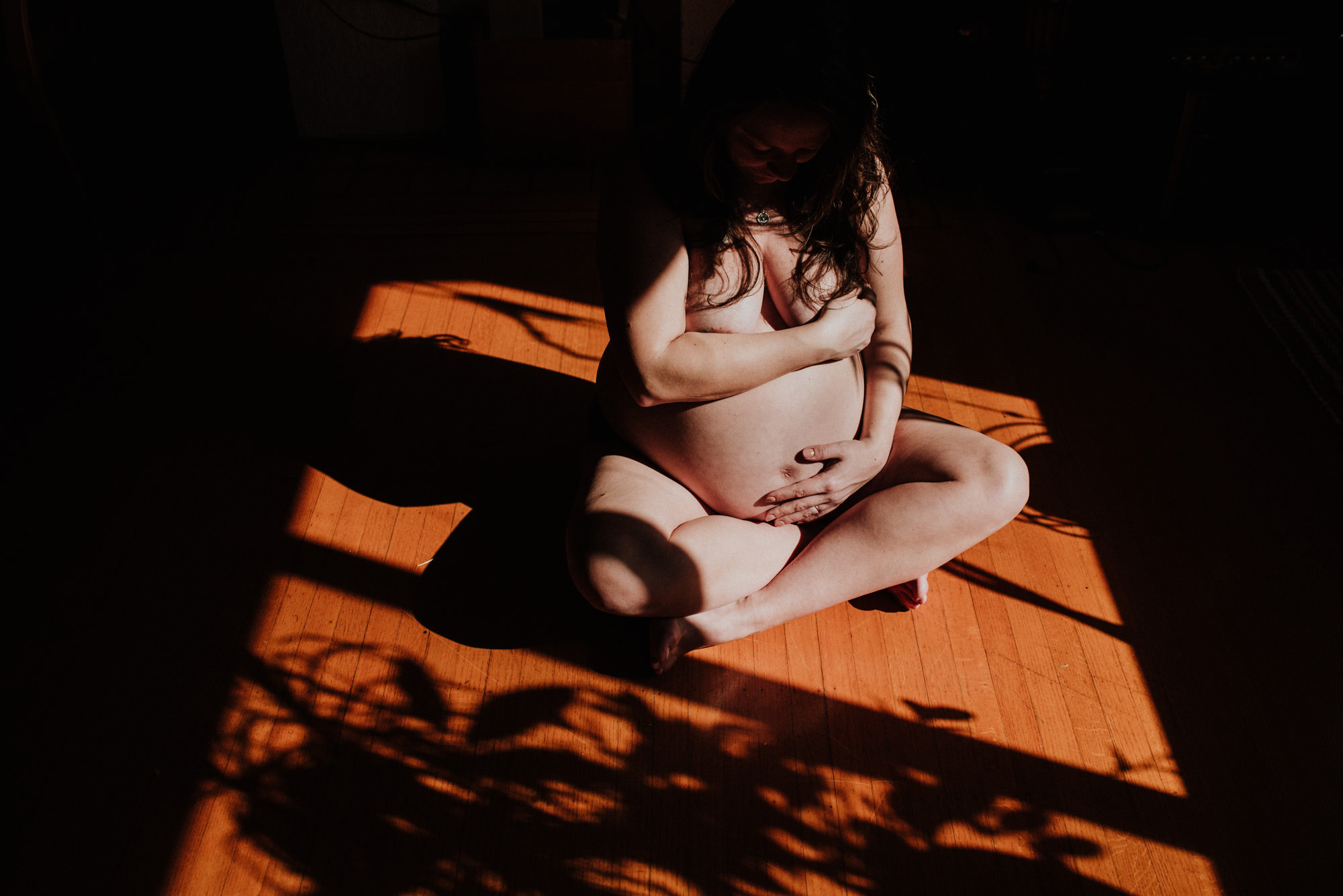 dark and moody maternity photo, photo of a pregnant woman sitting naked in sunshine